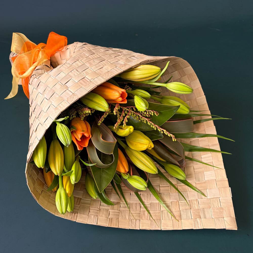 Sunset Lily Bouquet featuring orange and yellow Asiatic lilies with seasonal foliage wrapped in a woven flax-like mat